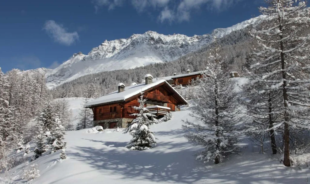 a wood and stone chalet is covered with snow under a high mountain ridge