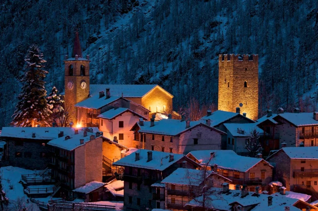 a church is lit up with lights at dusk, the snow-covered roofs of the village surrounding it