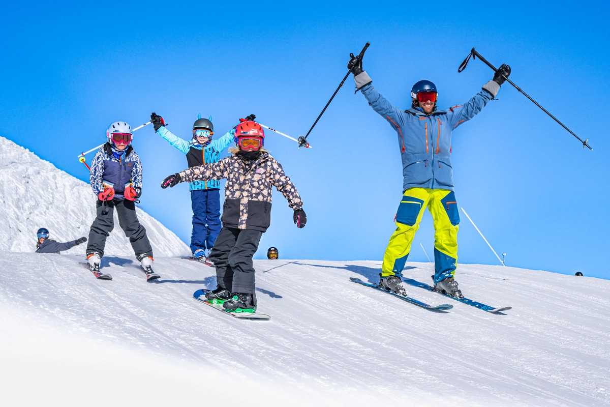 a family of skiers and snowboarder raise arms to camera as they ski past in a tight group