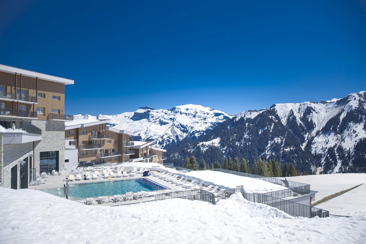 a mid-mountain situated alpine hotel exterior with a swimming pool surrounded by snow