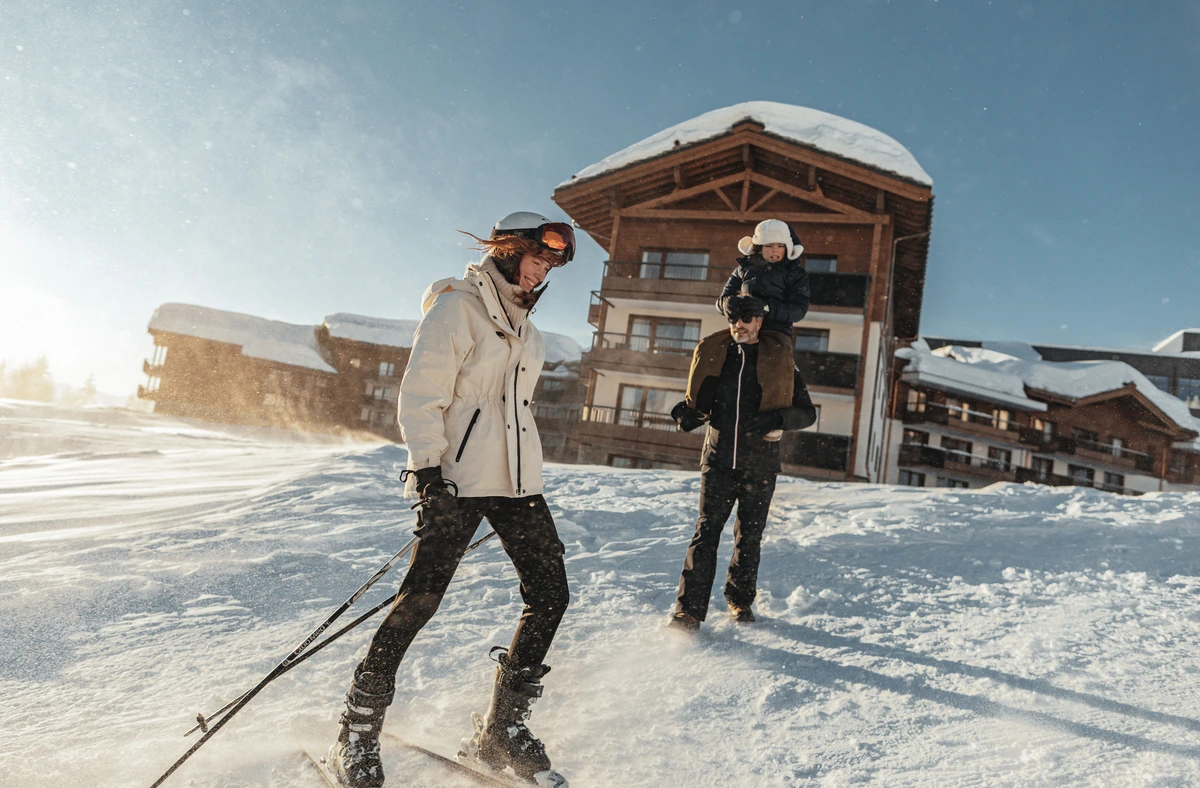 a dad carries a child on his shoulders, following a mum on skis, smiling, in front of a wooden alpine chalet-hotel next to the piste