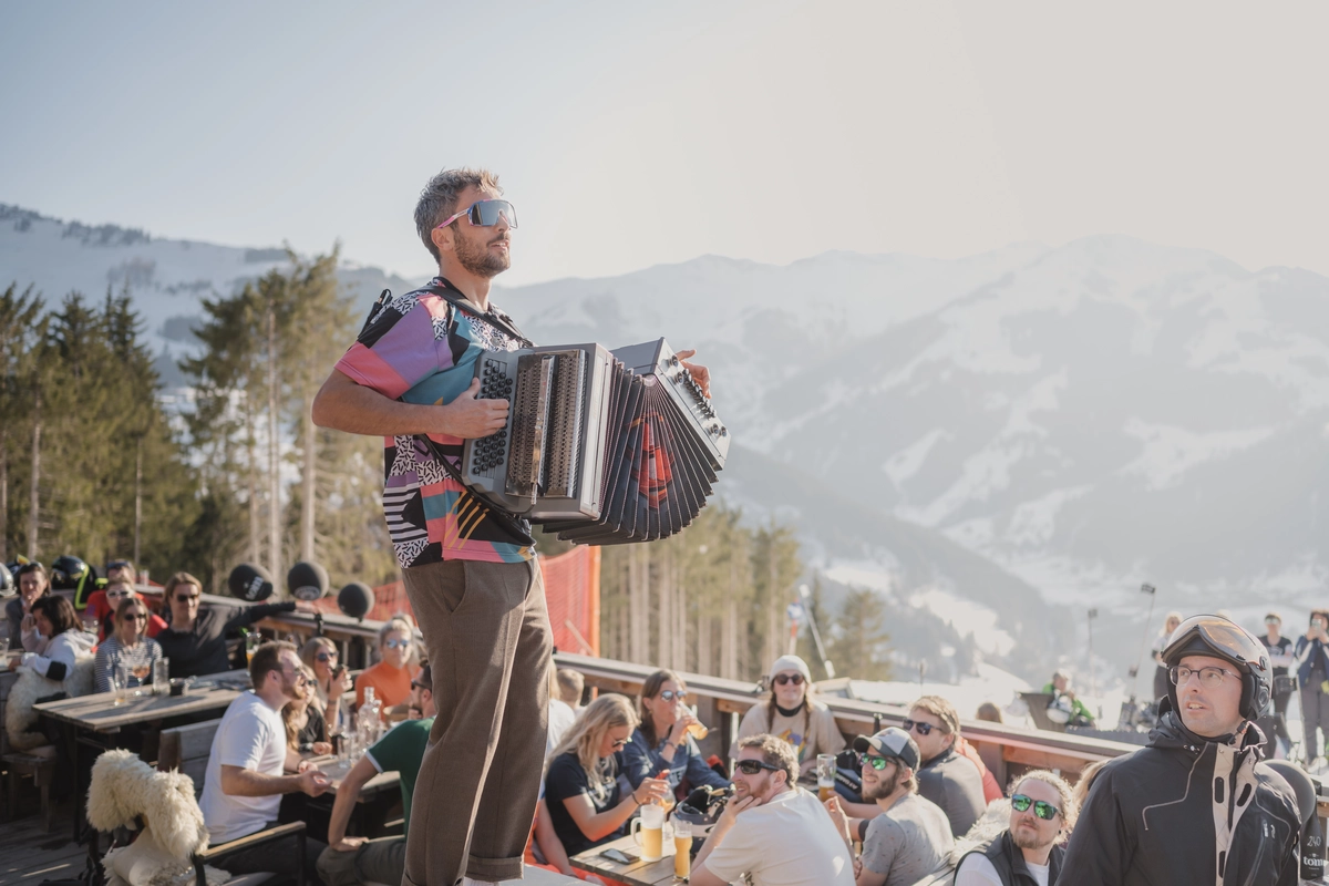 a man in a retro patterned t-shirt plays the accordion standing on a table of a busy ski terrace