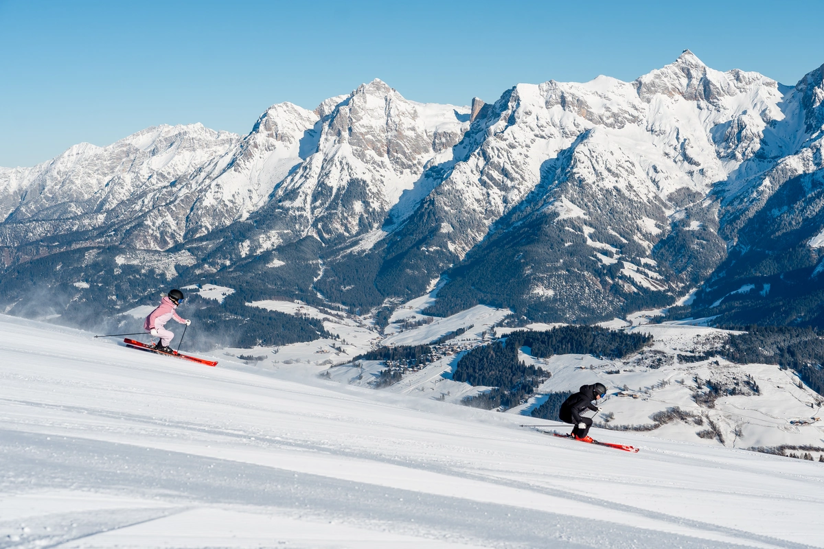 two skiers race down a groomed piste, behind them a snow-dusted massif