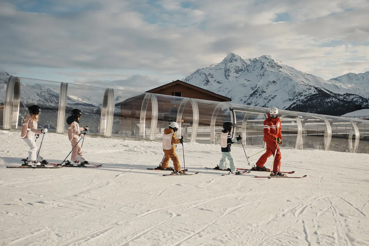 a line of kids in ski school follow an instructor in red down a nursery slope next to the magic-carpet tunnel