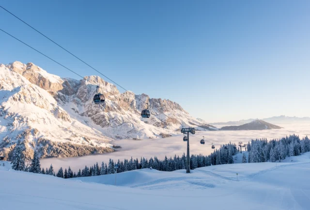 a snow dusted mountain ridge/massif behind a valley filled with clouds and a gondola travelling over snowy ski slopes