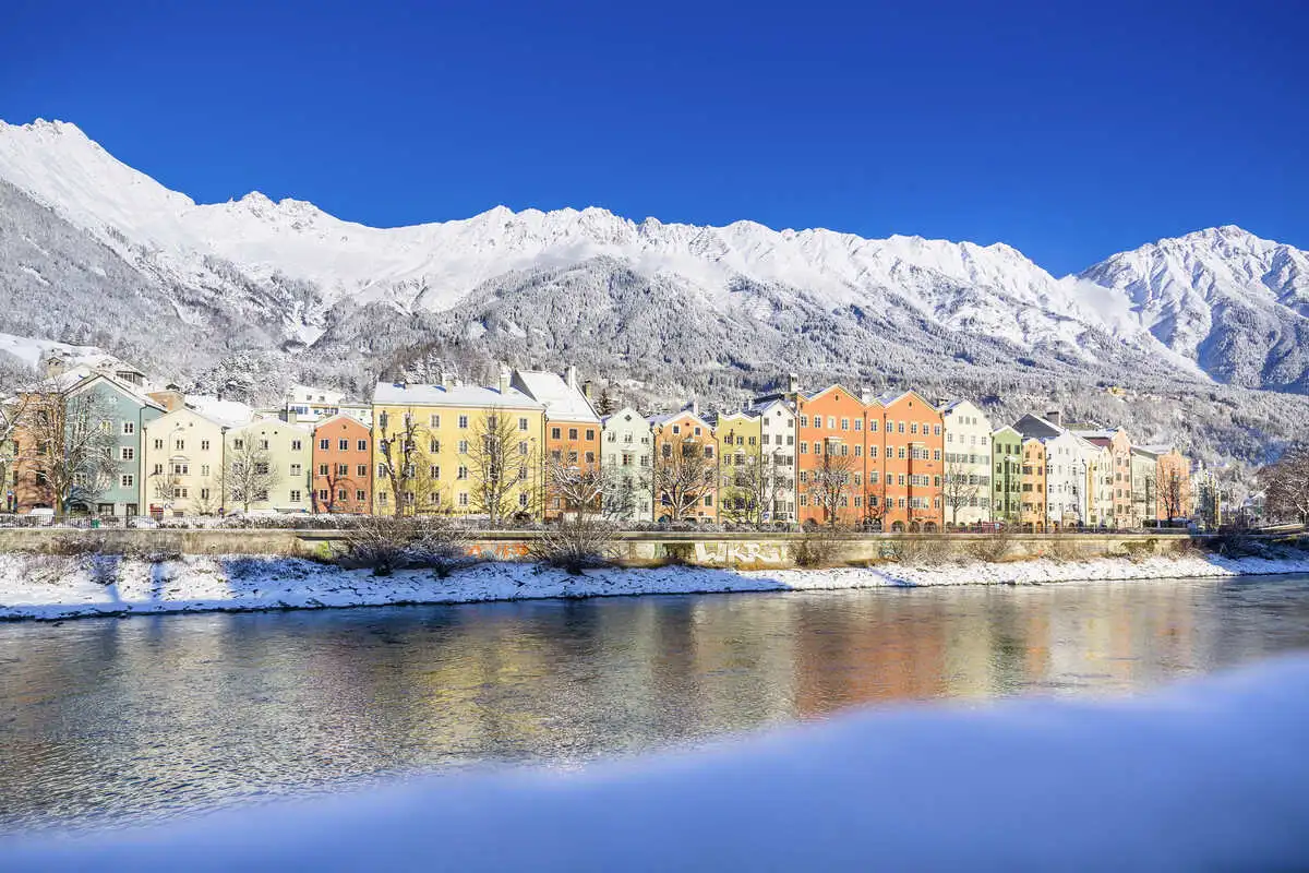 a colourful line of houses in front of a river, with snowy mountains as a backdrop