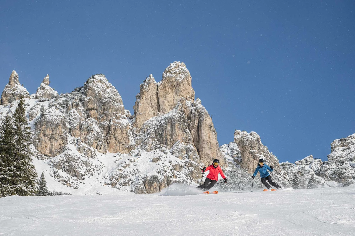 two skiers make turns on powdery piste in front of a Dolomite massif