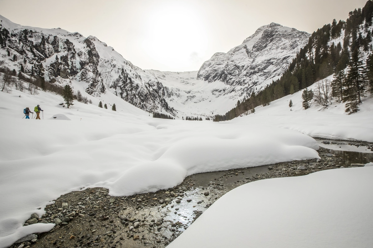 two snowshoers hike through a valley covered in snow besid a river | Winter hiking Lüsens-Fernerboden| © Innsbruck Tourismus / Erwin Haiden