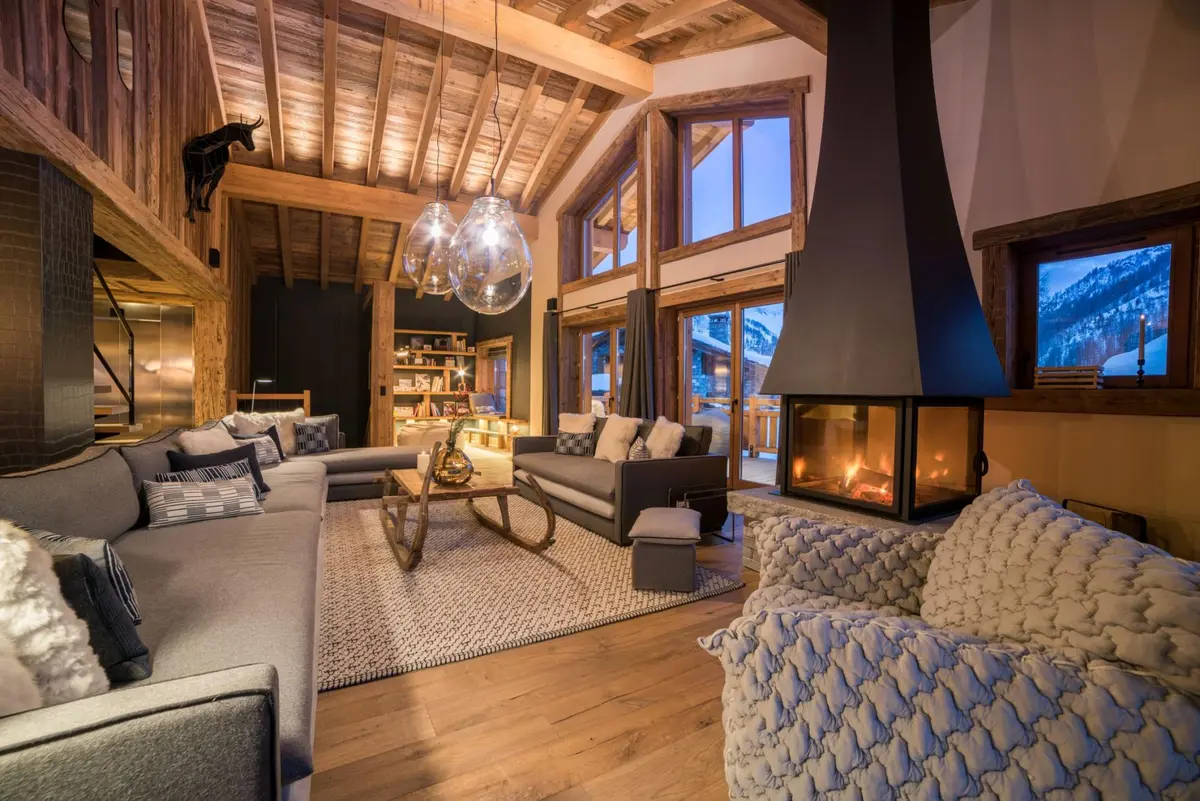 luxurious sitting room of a chalet pictured, with high vaulted ceiling, and a big fire in a modern fireplace